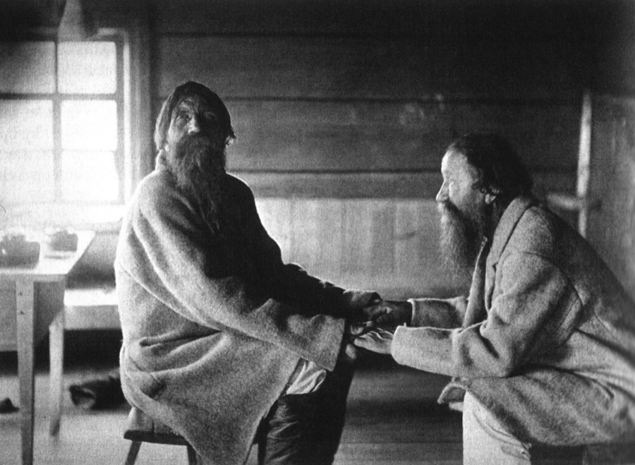 Photograph of two bearded Karelian men in a log-walled room, sitting face to face with their hands clasped between them