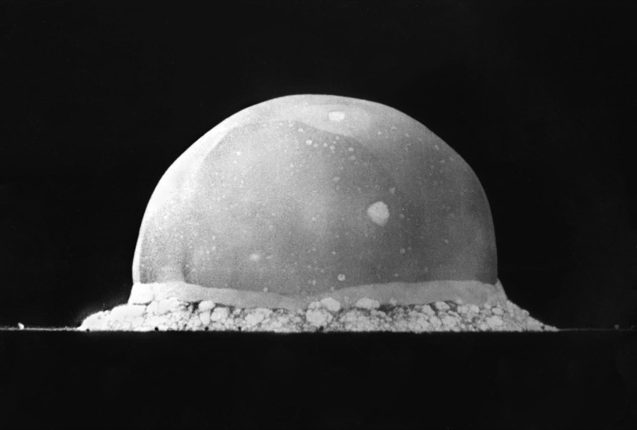 High-speed photograph of the Trinity test: a smooth white hemisphere of fire resting on the desert floor against a black sky